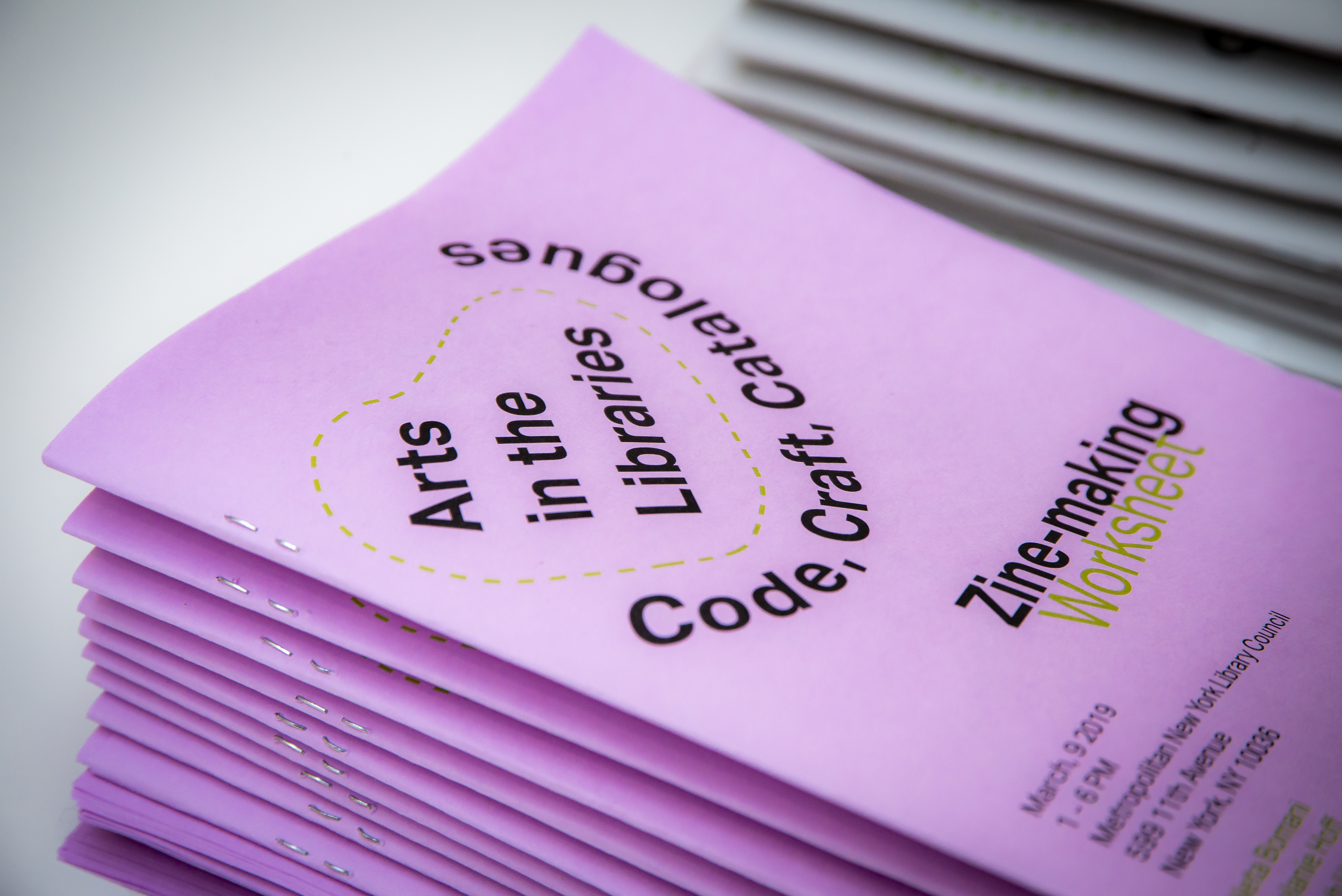 A close up of a stack of 10-15 worksheets printed on neon purple paper. The main title reads, “Arts in the Libraries” stylized in a black colored sans-serif font surrounded by a blob shape with a neon green dashed outline. The subtile reads, “Zine-making Worksheet.”
