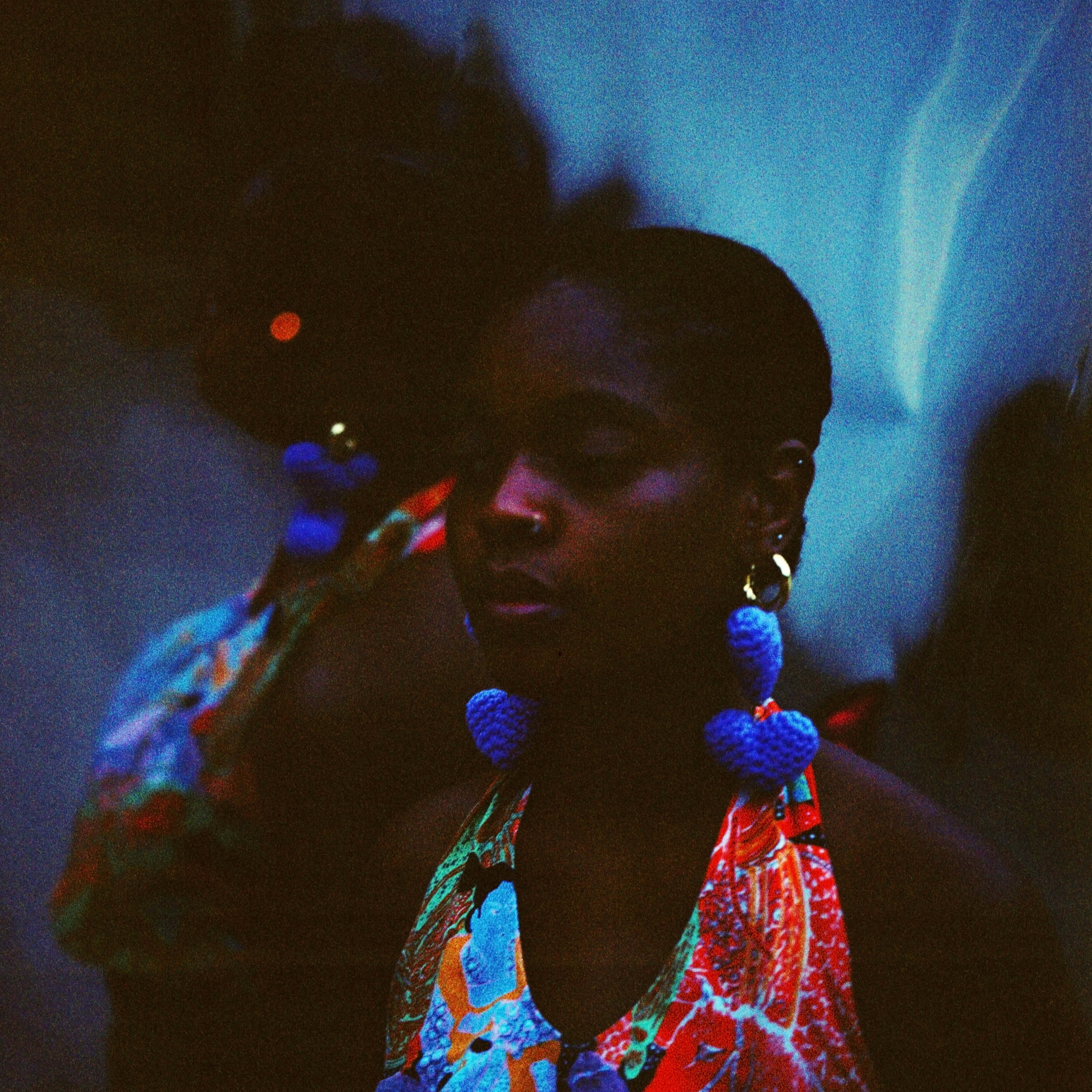 Neta, a deep brown skinned person, wears a colorful halter top and large lavender heart-shaped crochet earrings as she stands in front of a reflective backdrop that doubles and distorts the image of her profile..
