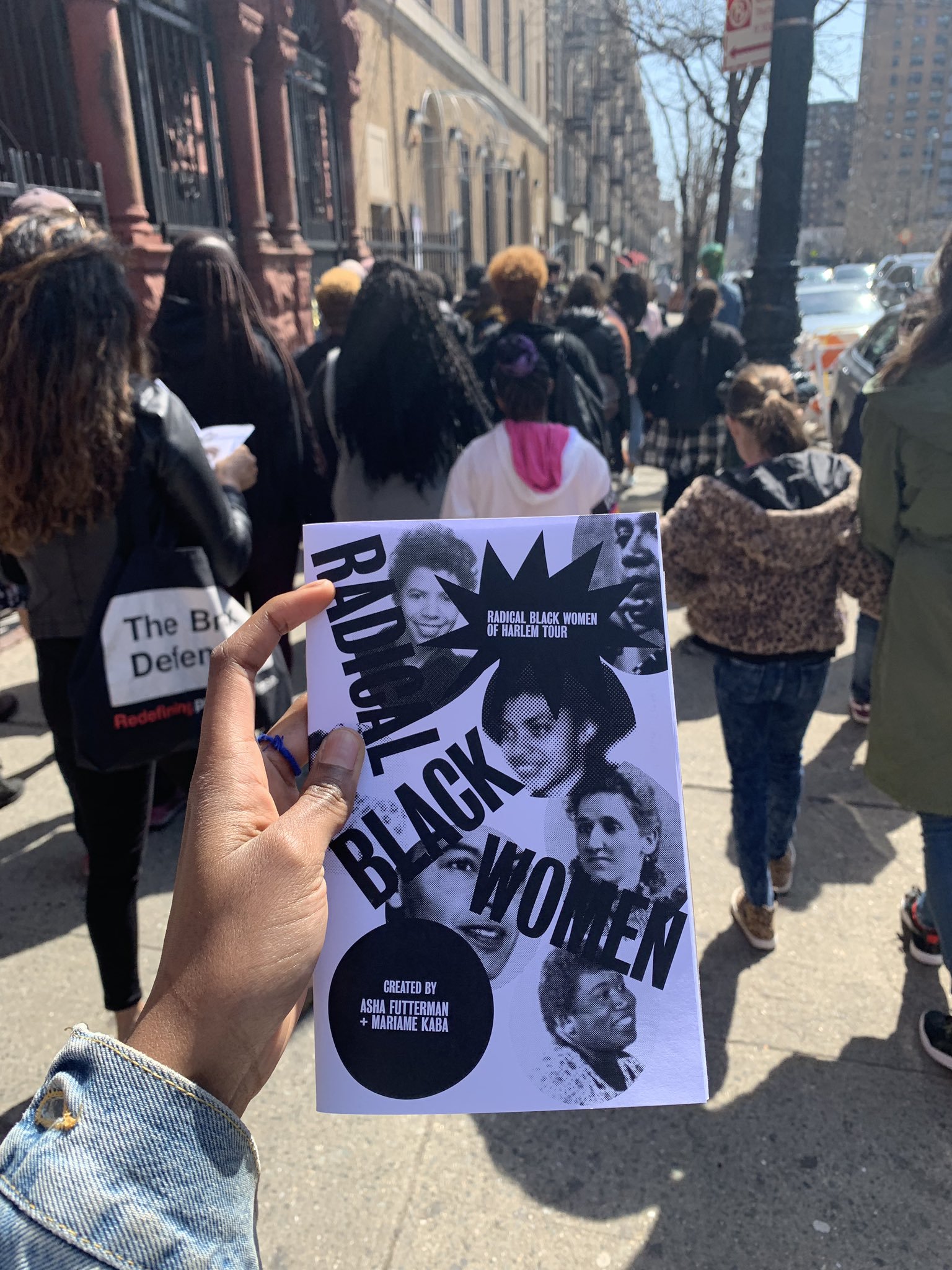 My hand holding the Radical Black Women of Harlem Tour guidebook in front of a crowd of people walking to the next stop on the tour.