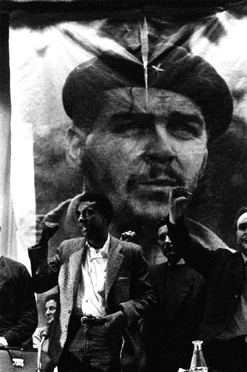 A black and white photograph of Kwame Ture standing in front of a poster of Che Guevara and raising a Black Power fist.