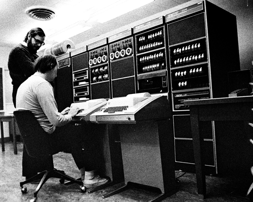 A black and white photograph of Unix developers Dennis Ritchie and Ken Thomas programming a unix machine.