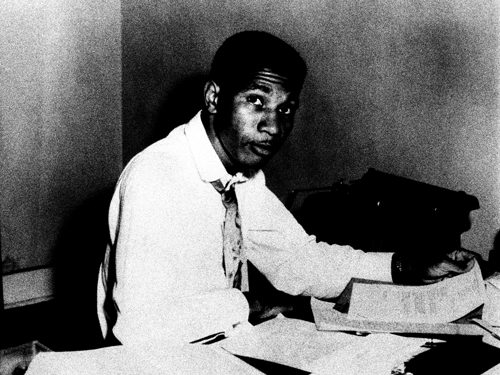 A black and white portrait of Medgar Evers sitting at a desk, wearing a white shirt and loose tie, thumbing through papers as he stares at the camera.